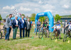 Innenminister Joachim Herrmann mit karierter Flagge und weitere Personen mit Blick zu 3 Bikern vor Parcour